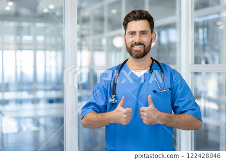 Smiling healthcare professional in blue scrubs giving thumbs up gesture, representing confidence and positivity in medical facility. Stethoscope around neck indicates medical role. Smiling healthcare professional in blue scrubs giving thumbs up gesture, representing confidence and positivity in medical facility. Stethoscope around neck indicates medical role. 122428946