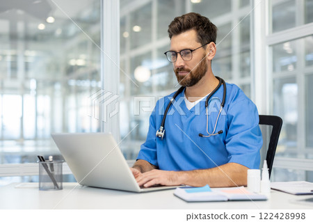 Man doctor in blue scrubs works on laptop in bright, modern medical office. Doctor wearing stethoscope, using technology for research and communication. Concept of healthcare, medicine, professional. Man doctor in blue scrubs works on laptop in bright, modern medical office. Doctor wearing stethoscope, using technology for research and communication. Concept of healthcare, medicine, professional. 122428998