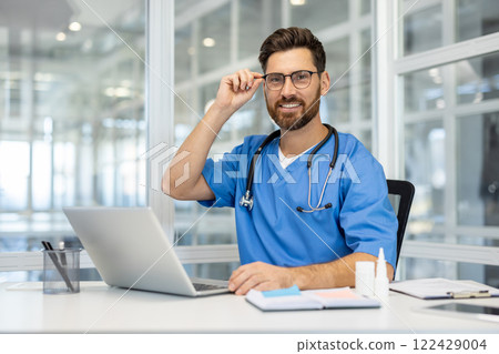 Smiling healthcare professional in blue medical scrubs at modern office with laptop. Stethoscope around neck, adjusting glasses, conveying professionalism, warmth, competence Smiling healthcare professional in blue medical scrubs at modern office with laptop. Stethoscope around neck, adjusting glasses, conveying professionalism, warmth, competence 122429004