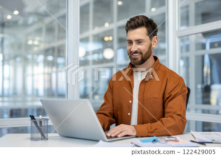 Smiling professional man using laptop in modern office. Confident worker enjoying work environment. Suitable for business, technology, productivity concepts. 122429005