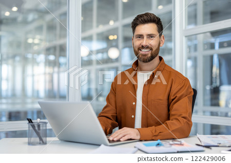 Confident professional man with a smile working on laptop in modern office setting, representing productivity and success at workplace. Workspace includes desk, papers, focus on technology. Confident professional man with a smile working on laptop in modern office setting, representing productivity and success at workplace. Workspace includes desk, papers, focus on technology. 122429006