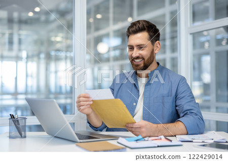 Man happily opening envelope at desk with laptop in modern office, illustrating positive emotion, communication, and work success. Bright setting with stylish decor. 122429014