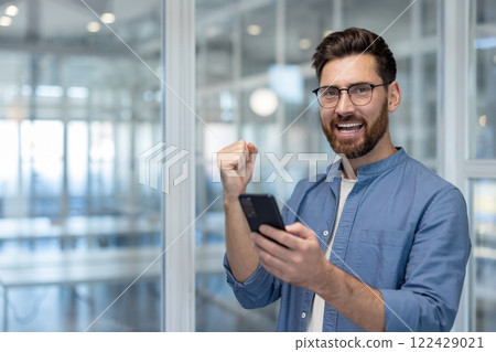 Portrait of a successful businessman at workplace inside office, a man with a phone in his hands celebrates victory and successful results of achievement, holds his hand up and looks at the camera. 122429021