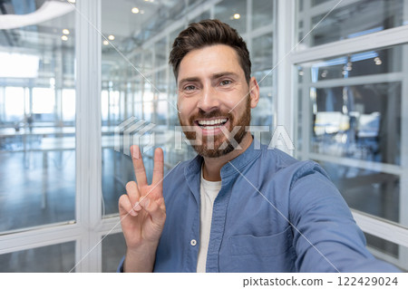 A cheerful man in a modern office setting takes a selfie, posing with a peace sign. The image conveys positivity, friendliness, and contemporary professional ambianc A cheerful man in a modern office setting takes a selfie, posing with a peace sign. The image conveys positivity, friendliness, and contemporary professional ambianc 122429024