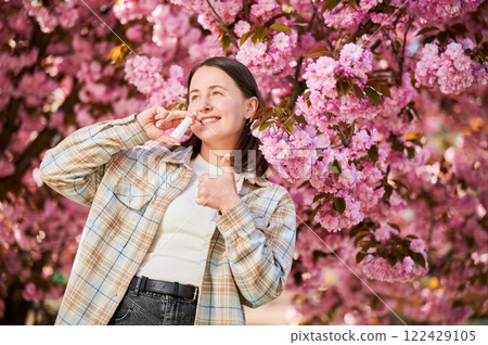 Woman allergic using medical nasal drops, suffering from seasonal allergy at spring in blossoming garden. Young woman treating runny nose in front of blooming tree outdoors. Spring allergy concept. Woman allergic using medical nasal drops, suffering from seasonal allergy at spring in blossoming garden. Young woman treating runny nose in front of blooming tree outdoors. Spring allergy concept. 122429105