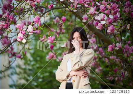 Woman allergic suffering from seasonal allergy at spring in blossoming garden at springtime. Young woman sneezing, closing nose by hand in front of blooming tree. Spring allergy concept 122429115