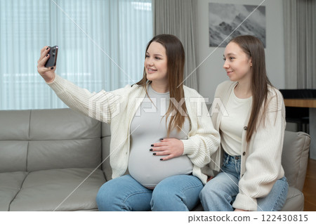 Young pregnant woman taking a selfie with her female friend at home on a sofa, smiling, with copy space. Concept of friendship, maternity, capturing special memories during pregnancy. 122430815