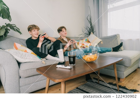 Two teenager boys are focused on their video game consoles while sitting on couch at home. Brothers Gamers enjoying Playing Video Games indoors. Siblings Spending time at home with snacks and drinks. Two teenager boys are focused on their video game consoles while sitting on couch at home. Brothers Gamers enjoying Playing Video Games indoors. Siblings Spending time at home with snacks and drinks. 122430902
