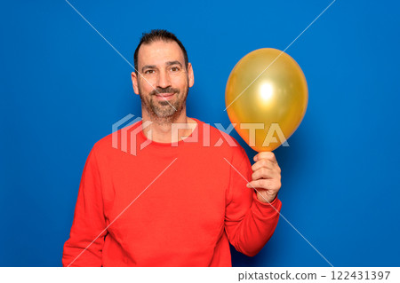 Bearded hispanic man in his 40s wearing a red sweater holding an orange balloon filled with happiness and satisfaction. Isolated on blue studio background Bearded hispanic man in his 40s wearing a red sweater holding an orange balloon filled with happiness and satisfaction. Isolated on blue studio background 122431397