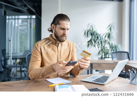 Focused man sitting at a desk in an office environment interacting with a smartphone and a credit card, implying online payment, banking, or identity verification. 122431666