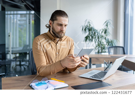 A professional man sits at a desk in a modern office, holding a tablet and focusing on work. His setup includes a laptop, notebook, and accessories indicative of productivity. A professional man sits at a desk in a modern office, holding a tablet and focusing on work. His setup includes a laptop, notebook, and accessories indicative of productivity. 122431677