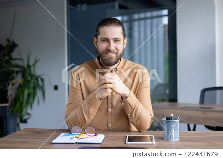 Businessman smiling at desk in a modern office setting, showing a cheerful demeanor while seated near a tablet and office supplies, conveying professionalism and approachability Businessman smiling at desk in a modern office setting, showing a cheerful demeanor while seated near a tablet and office supplies, conveying professionalism and approachability 122431679