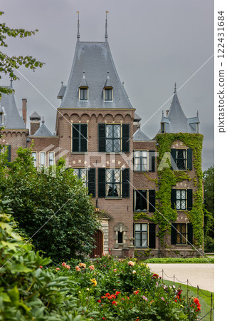 A picturesque view of a historic brick mansion with a round tower, pointed roof, and arched windows, surrounded by lush greenery, manicured gardens, and a peaceful, cloudy sky. A picturesque view of a historic brick mansion with a round tower, pointed roof, and arched windows, surrounded by lush greenery, manicured gardens, and a peaceful, cloudy sky. 122431684