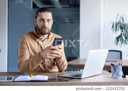 A professional individual sitting at a desk with a laptop, using a smartphone, creating a productive working environment. A professional individual sitting at a desk with a laptop, using a smartphone, creating a productive working environment. 122431702