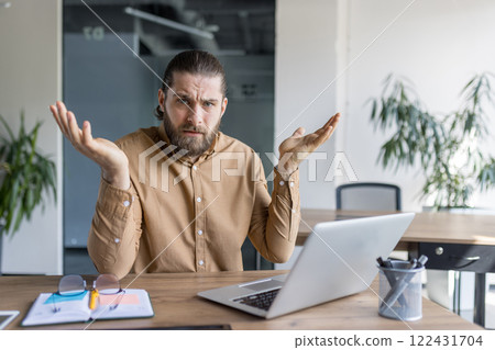 A man with a puzzled expression sits at a desk using a laptop, gesturing with raised arms, conveying confusion or hesitation while working in an office or professional setting. 122431704
