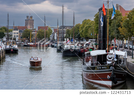 Dordrecht, South Holland, Holland - 09.07.2024: Serene harbor with moored boats under a dramatic cloudy sky. Quaint historic buildings and trees line the waterfront, reflecting in the calm green water Dordrecht, South Holland, Holland - 09.07.2024: Serene harbor with moored boats under a dramatic cloudy sky. Quaint historic buildings and trees line the waterfront, reflecting in the calm green water 122431713