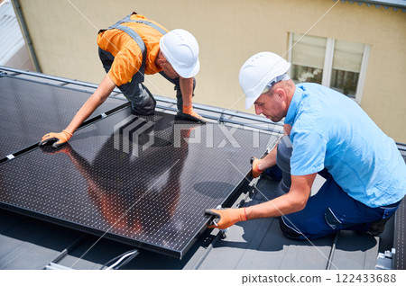 Workers building solar panel system on rooftop of house. Two men installers in helmets installing photovoltaic solar module outdoors. Alternative, green and renewable energy generation concept. 122433688