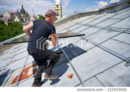Workers building solar panel system on metal rooftop in urban setting. Two men installers carrying photovoltaic solar module outdoors. Alternative, green and renewable energy generation concept. 122433692