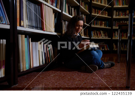 Woman reading a book seated on the floor of a library 122435139