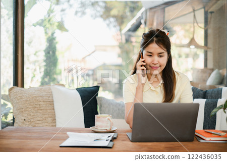A young woman multitasks with a laptop and smartphone in a bright, modern living room, showcasing a comfortable remote work environment. 122436035