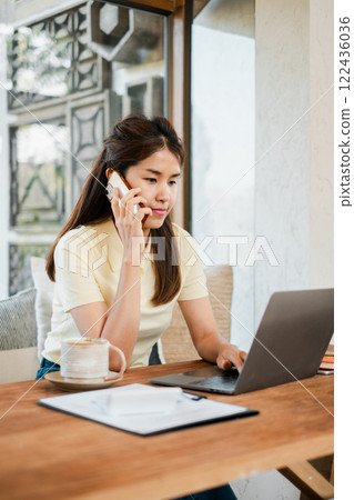 Young woman multitasking at home office, using a laptop and smartphone, with coffee and documents on a wooden desk. 122436036