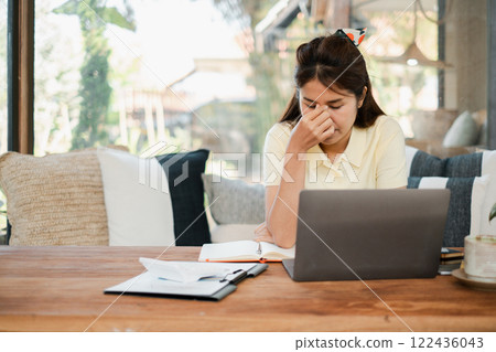 Woman feeling stressed while working from home, sitting at a wooden table with a laptop and documents in a cozy living room. Woman feeling stressed while working from home, sitting at a wooden table with a laptop and documents in a cozy living room. 122436043