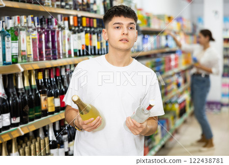 Young male customer deciding on buying bottle of wine or vodka Young male customer deciding on buying bottle of wine or vodka 122438781