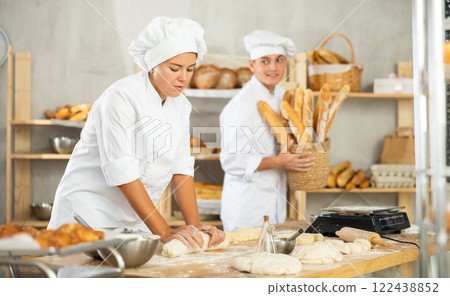 Young female baker kneading dough and guy carrying baguettes Young female baker kneading dough and guy carrying baguettes 122438852