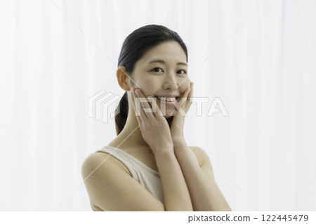 A smiling young woman wearing a tank top on a white background and holding both hands on her cheeks Beauty 122445479