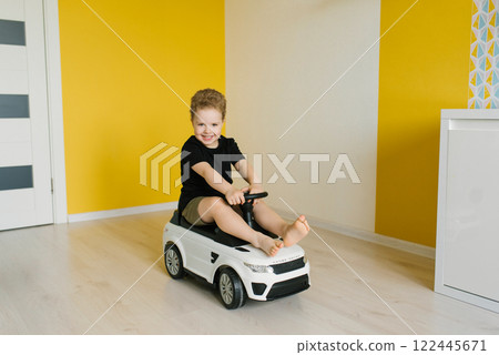 Child boy is sitting in toy car, smiling and enjoying himself in room is brightly lit and has yellow wall, creating cheerful atmosphere 122445671