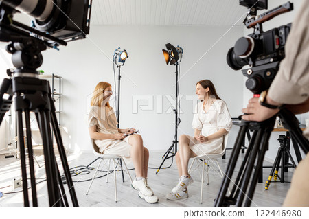 Interview set. Friendly dialogue. Two smiling girls sit chairs illuminated video studio. Female models posing business concept photo. 122446980