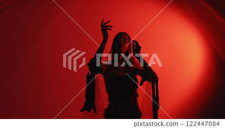 Spiritual dance. Soul healing. Relaxed woman silhouette in trance wearing polyethylene rags performing graceful hand movements isolated on dark red glow copy space background. Spiritual dance. Soul healing. Relaxed woman silhouette in trance wearing polyethylene rags performing graceful hand movements isolated on dark red glow copy space background. 122447084