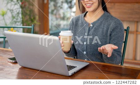 A smiling Asian woman discusses work through an online video meeting, working remotely from a cafe. 122447282