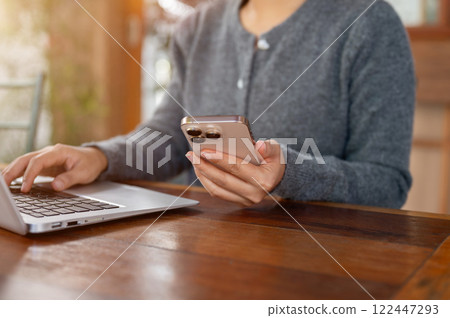 A close-up of a happy Asian woman reads messages on her smartphone while working on her laptop. A close-up of a happy Asian woman reads messages on her smartphone while working on her laptop. 122447293