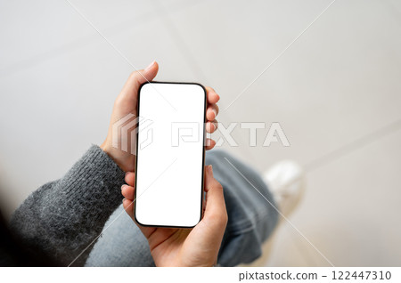 A top view of a woman holds a smartphone with a white screen mockup while sitting indoors. 122447310