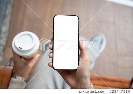 A top view of a smartphone mockup in a woman's hand. A woman holds a coffee cup and a smartphone. 122447919