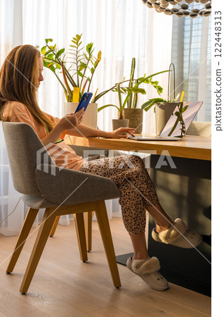 Woman working from home office with smartphone and laptop in a cozy, sunlit in homewear 122448313