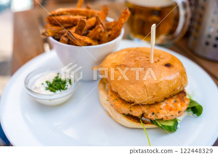 Close-up of a gourmet salmon burger served with crispy sweet potato fries and herb dip, beer. 122448329