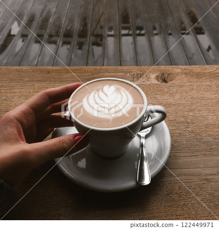 White coffee cup stands on wooden table on cafe terrace on a city street. Morning coffee outdoors in rainy day. Cappuccino, latte cup in female hand. White coffee cup stands on wooden table on cafe terrace on a city street. Morning coffee outdoors in rainy day. Cappuccino, latte cup in female hand. 122449971