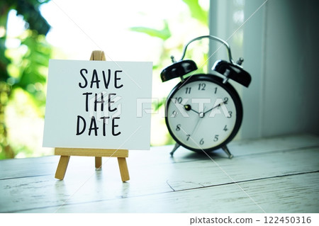 Save the Date quotes text on paper card with easel and alarm clock in the window background 122450316