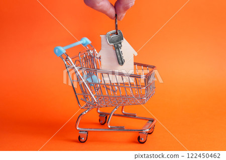 Hand places bunch of keys with wooden house keychain into mini shopping cart. Bright orange background. Side view. Real estate and home ownership concept Hand places bunch of keys with wooden house keychain into mini shopping cart. Bright orange background. Side view. Real estate and home ownership concept 122450462