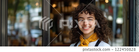 Smiling young woman hotel staff member with curly hair and yellow scarf standing at the entrance of a European hospitality establishment. Front view 122450828