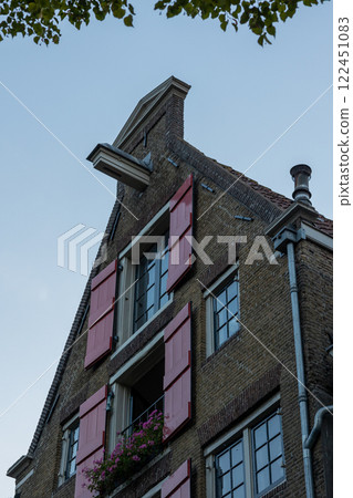 A historic brick building with red wooden shutters and large windows. A small balcony with pink flowers adds charm. The stepped gable and pulley beam hint at Dutch architectural style. A historic brick building with red wooden shutters and large windows. A small balcony with pink flowers adds charm. The stepped gable and pulley beam hint at Dutch architectural style. 122451083