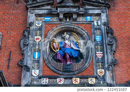 A historic architectural relief on a red brick wall features a statue of a seated woman in blue robes holding a shield and a golden palm leaf, surrounded by ornate stonework and heraldic crests. 122451087