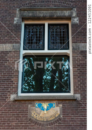 A brick building facade with a decorative window featuring stained glass. Below, a sundial with golden details and a blue emblem displays the Dutch phrase: "I count only the sunny hours". A brick building facade with a decorative window featuring stained glass. Below, a sundial with golden details and a blue emblem displays the Dutch phrase: "I count only the sunny hours". 122451091