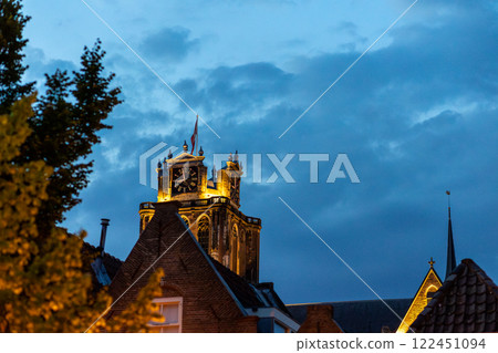 An illuminated historic clock tower rises above the rooftops at dusk, with blue sky in the background. Warm lights highlight the architectural details, creating contrast with the evening sky. An illuminated historic clock tower rises above the rooftops at dusk, with blue sky in the background. Warm lights highlight the architectural details, creating contrast with the evening sky. 122451094