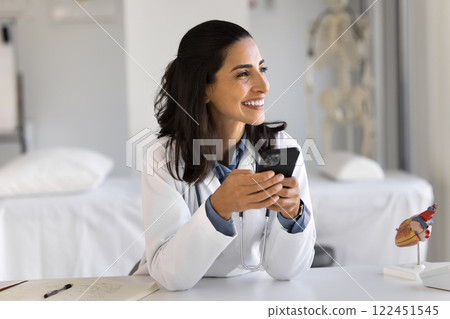 Cheerful young lady doctor sit at work table hold cellphone 122451545