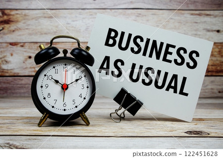 Business as Usual text on paper card with alarm clock on wooden background 122451628