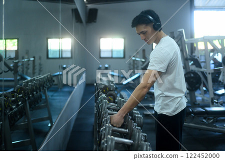 Man wearing headphones picking weights from a gym rack in front of mirror 122452000