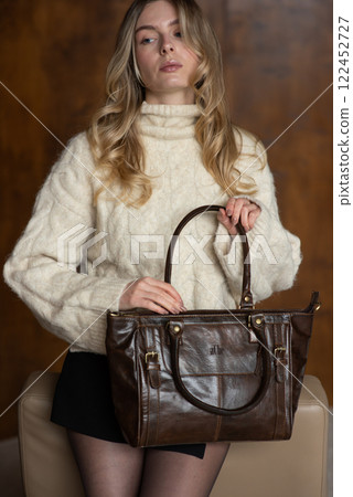 A girl in a warm sweater, a black short skirt, nylon tights is posing with a brown leather bag. 122452727